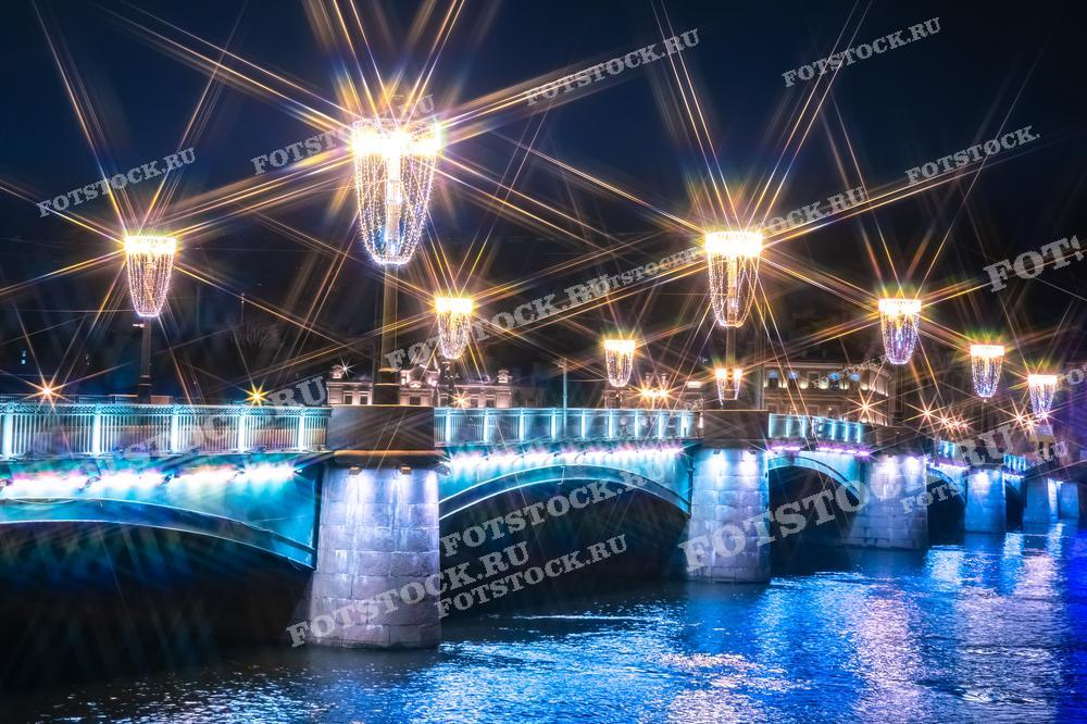 Фотография представляет собой вид на мост в Санкт-Петербурге, украшенный праздничной иллюминацией. Игра света на воде и звездообразные блики от фонарей создают ощущение праздника и волшебства. Этот снимок идеален для иллюстрации новогоднего или рождественского настроения в городе и может быть использован в качестве открытки, в туристических проспектах или как декоративный элемент в оформлении мероприятий.